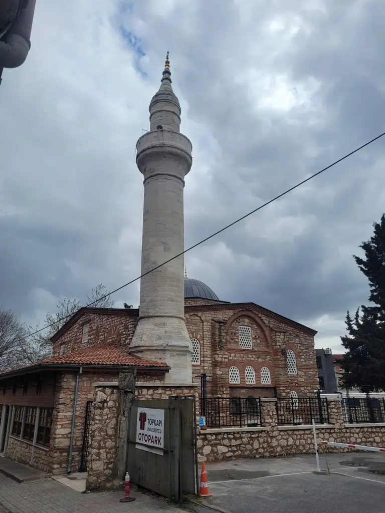 Atik Mustafa Paşa Camii'nin sokaktan görünüşü; önde gri kesme taş minare, arkada tuğla gövde ile kurşun kubbe, solda kiremit çatılı ahşap son cemaat yeri ve bulutlu gök.