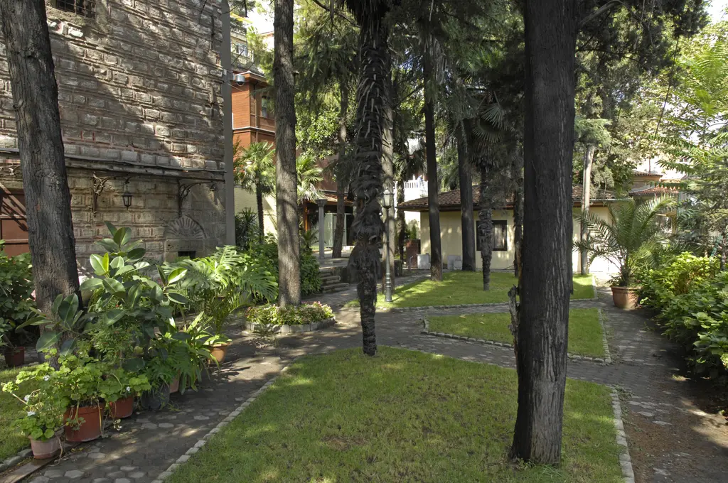 Patrikhane bahçesi; yüksek şemsiye çamlar, çakıllı patika, sıralanmış saksılar ve arkada yerleşkenin ahşap kanadı.