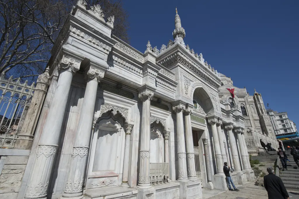Pertevniyal Valide Sultan Camii'nin anıtsal avlu kapısı; çifte sütunlar, mermer cephe, yanda merdiven ve kapıya yürüyen insanlar.