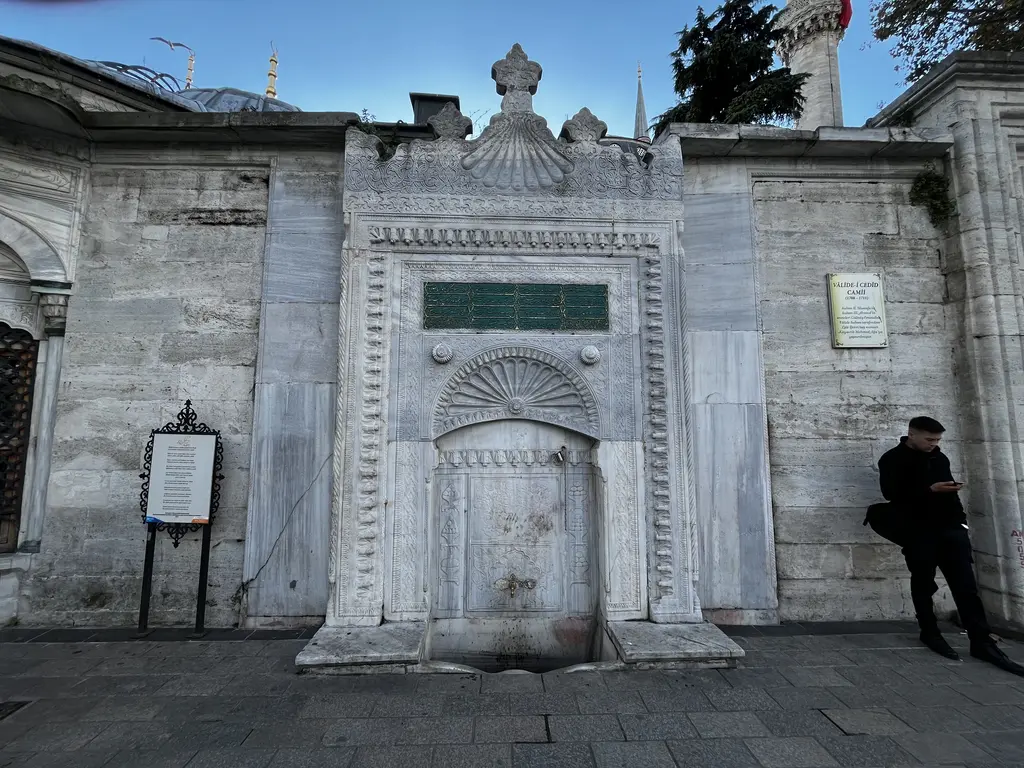 Yeni Valide Külliyesi'nin çeşmesi; oymalı mermer cephe, yazı levhası, kemerli niş ve önünde duran bir adam.