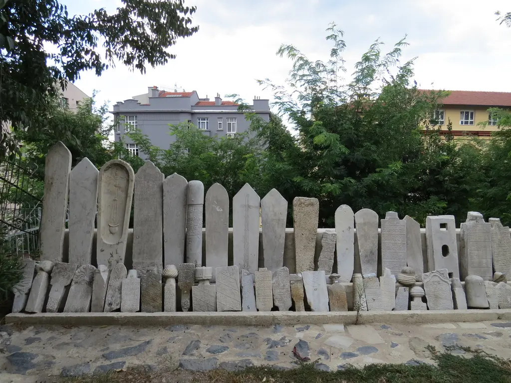 Hazirenin kenarında duvar boyunca yan yana dizilmiş eski mezar taşları; arkada yeşillik ve binalar.