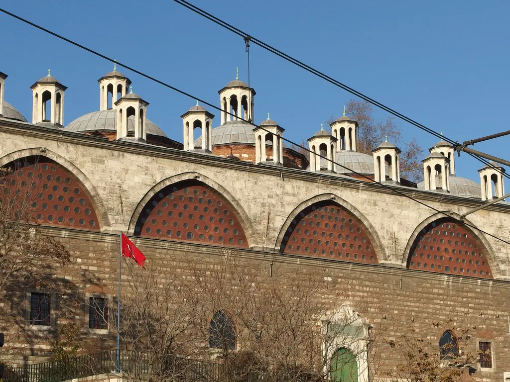 Tophane-i Âmire'de yan yana dizilen küçük fenerli kubbeler; altında kemerli taş cephe ve bir bayrak.
