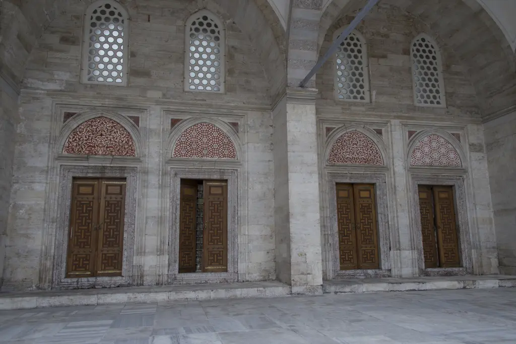 Şehzade Camii'nin avlu cephesi; yan yana dört ahşap kapı, her birinin üstünde kırmızı geometrik kafes alınlık, üst katta demir parmaklıklı pencereler.