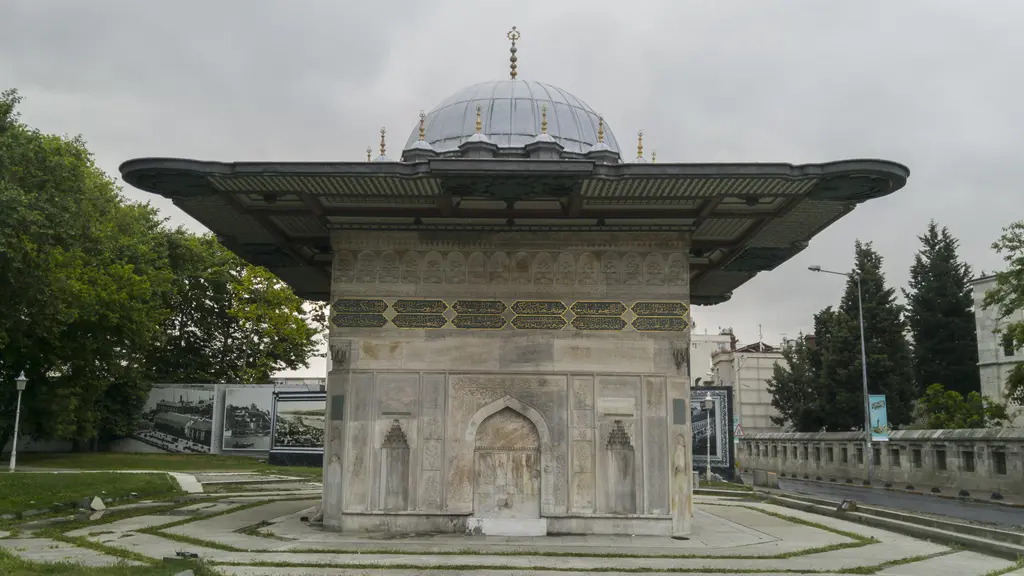 Caddeden Tophane Çeşmesi; mavi gökyüzü altında kare gövde ve geniş saçak, arkada caminin kubbeleri ve minaresi, önde boş yol.
