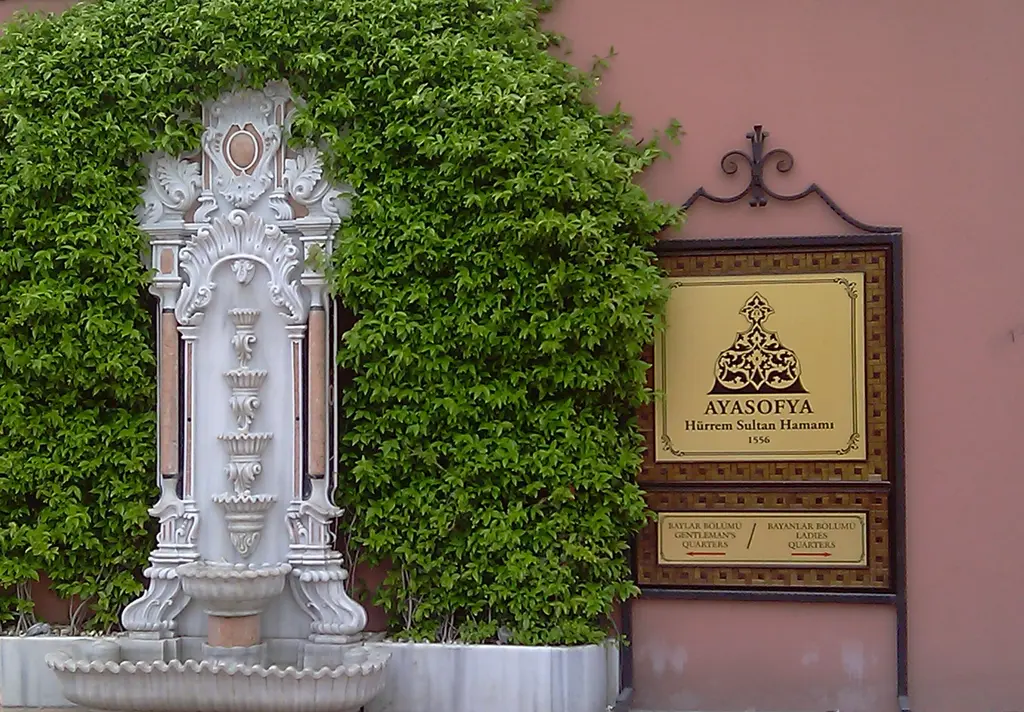 Pembe duvarda sarmaşığın çevrelediği beyaz mermer çeşme ve yanında "Ayasofya Hürrem Sultan Hamamı 1556" yazan altın renkli levha.