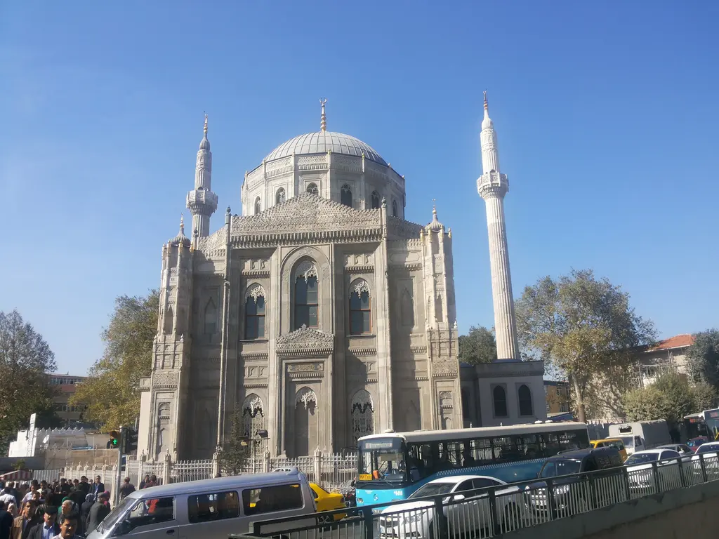 Pertevniyal Valide Sultan Camii; mavi gökyüzü altında taş cephe, kubbe, iki minare ve önde caddede duran otobüsler.