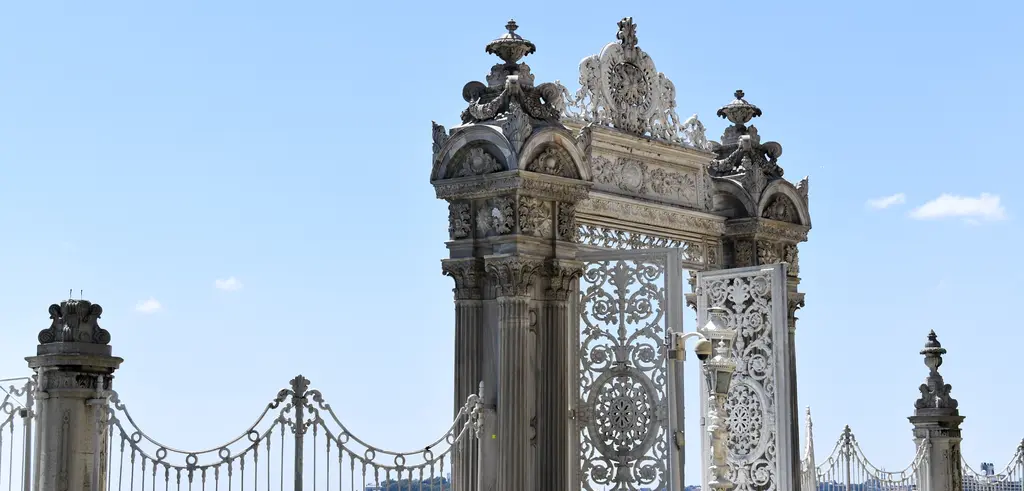 Dolmabahçe Sarayı'nın deniz kapısı; yandan bakışta oymalı taş ayaklar, süslü demir kanatlar ve arkada açık mavi gökyüzü.