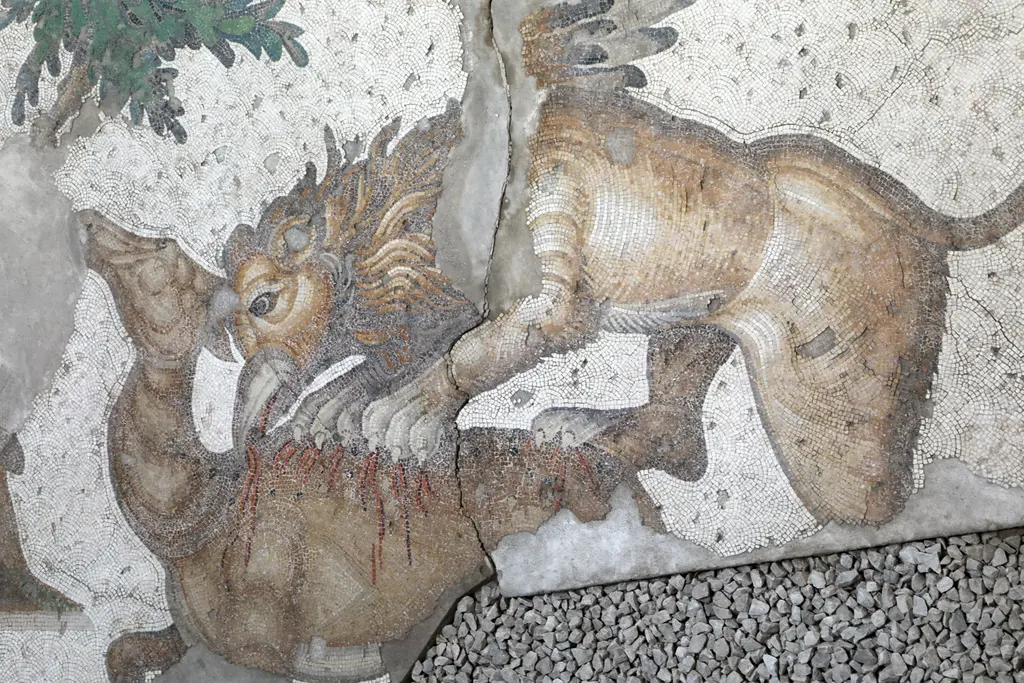 Mozaikte kanatlı bir yaratık bir ceylanın boynunu ısırıyor; ceylan dizleri üzerine çökmüş, arkada bir ağaç.
