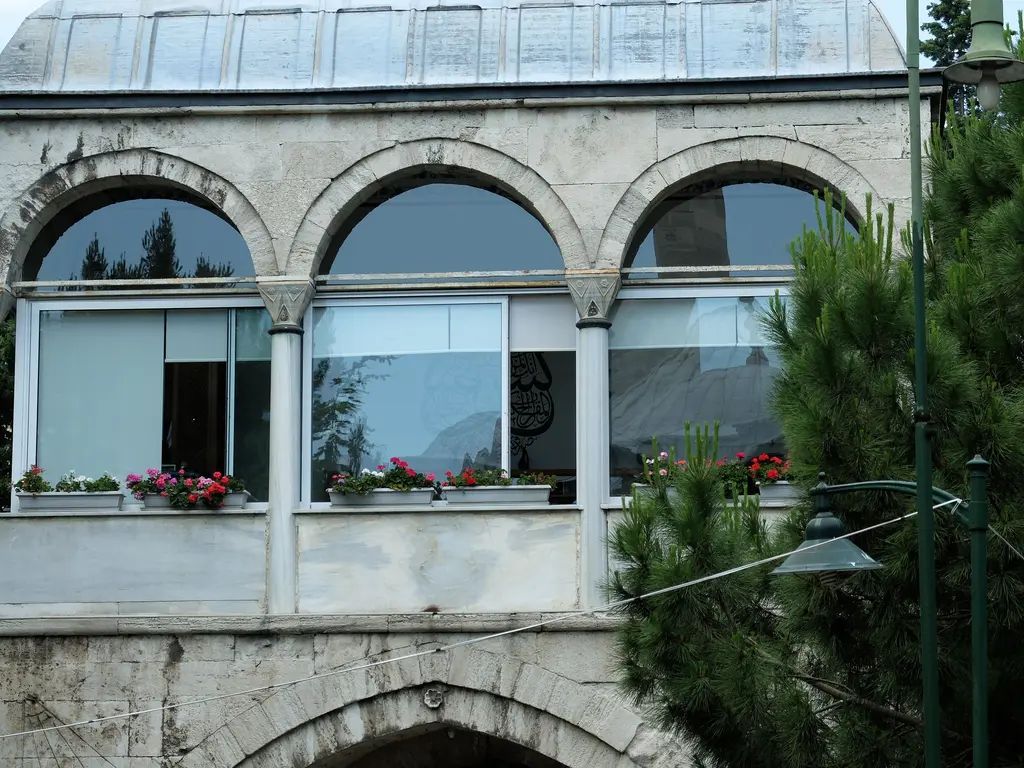 Kütüphane binası; sütunlara oturan yuvarlak kemerli balkon, üstte dikdörtgen pencereler, önünde merdiven ve saksı çiçekleri.