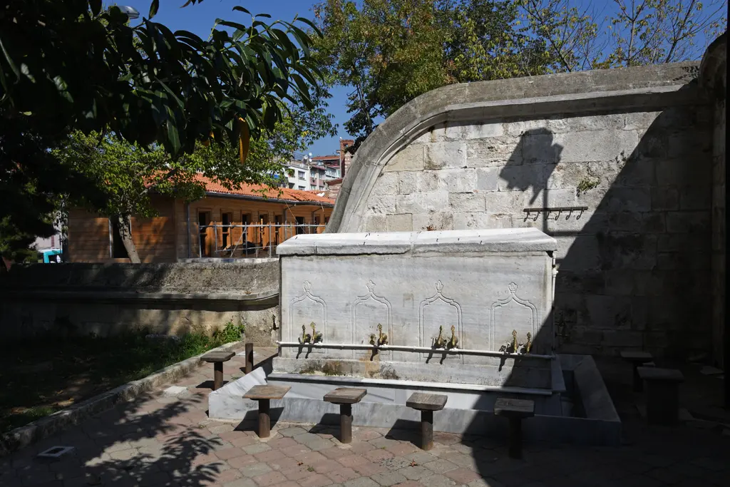 Selimiye Camii avlusunda mermer çeşme; uzun mermer hazne, sıra musluklar, önünde taş tabureler ve arkada taş duvar.