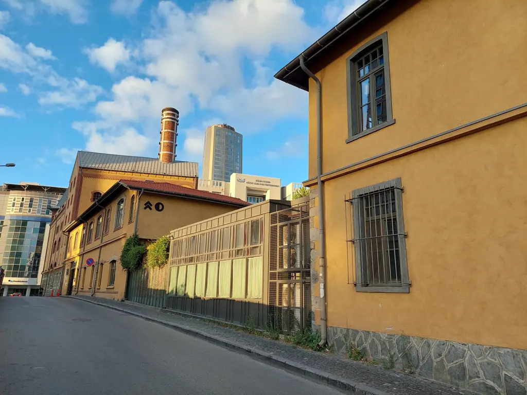 Bomonti'de eski bira fabrikasının sarı sıvalı tarihî binaları ve tuğla bacası, önde eğimli bir sokak, arkada modern bir kule.
