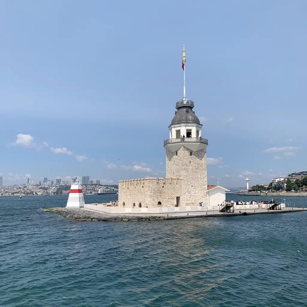 Kız Kulesi; yakından taş kaide, tepede bayraklı kule, iki yanda küçük fenerler ve mavi deniz.