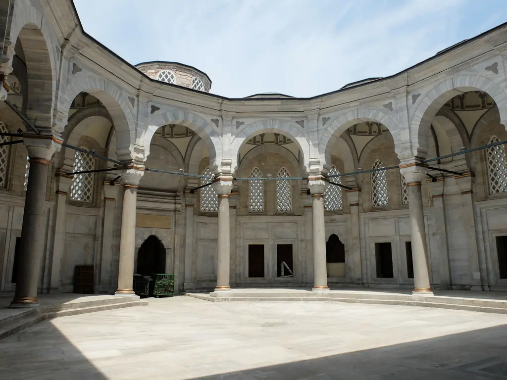 Nuruosmaniye Camii'nin avlusu; yarım daire çizerek uzanan revak dizisi, sıra sıra mermer sütunlar ile yuvarlak kemerler, açık renk taş zemin ve parlak gökyüzü.
