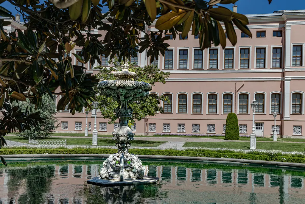 Dolmabahçe Sarayı'nın Harem bahçesi; ağaç dallarının altından görülen havuz, ortada fıskiye ve arkada pembe cepheli bina.