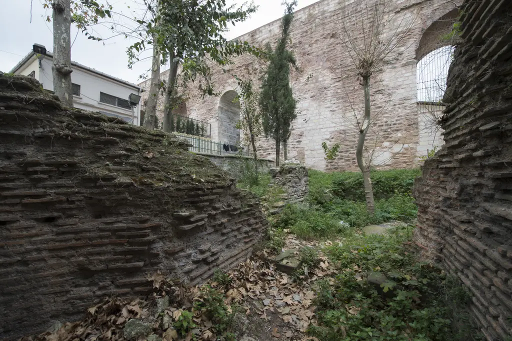 Bozdoğan Kemeri'nin otlarla kaplı eteği; önde kırık tuğla sıraları ve moloz taş dolgu, yabani otlar ile yaprak döküntüsü, arkada kemerin gövdesi ve ince selviler.
