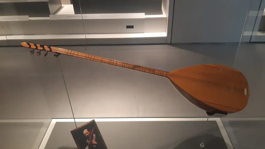 AKM Müzik Platformu'nda bir vitrinde uzun saplı bağlama; açık renkli ahşap gövde, ince sap ve altta bir portre fotoğrafı.