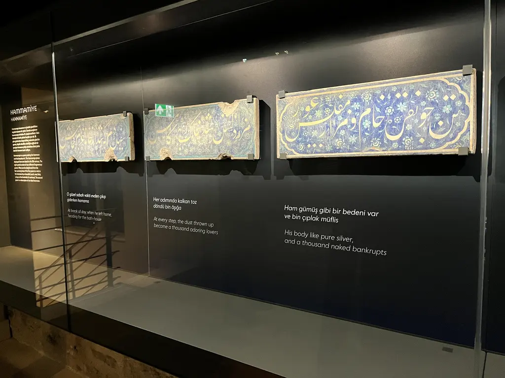 Çinili Hamam Müzesi'nde vitrin; üç büyük İznik çini levhası, lacivert zemin üzerine beyaz hatla yazı, yanında Türkçe ve İngilizce metinler.