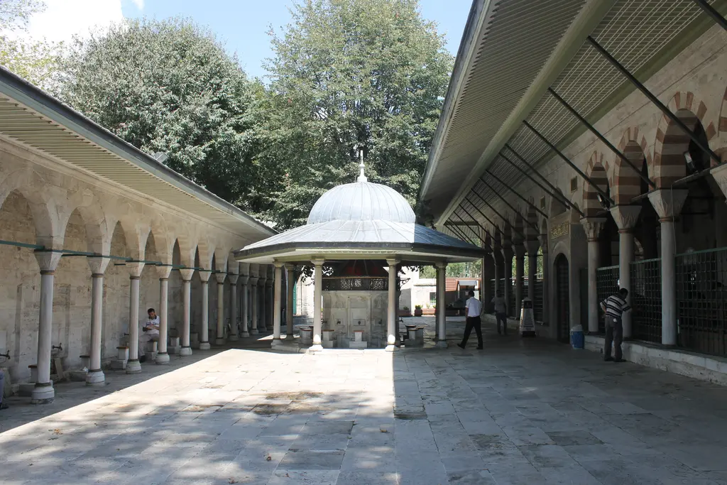 Kılıç Ali Paşa Camii'nde avlu; ortada kubbeli şadırvan, iki yanda sütunlu revaklar, taş döşeme ve arkada yeşil ağaçlar.
