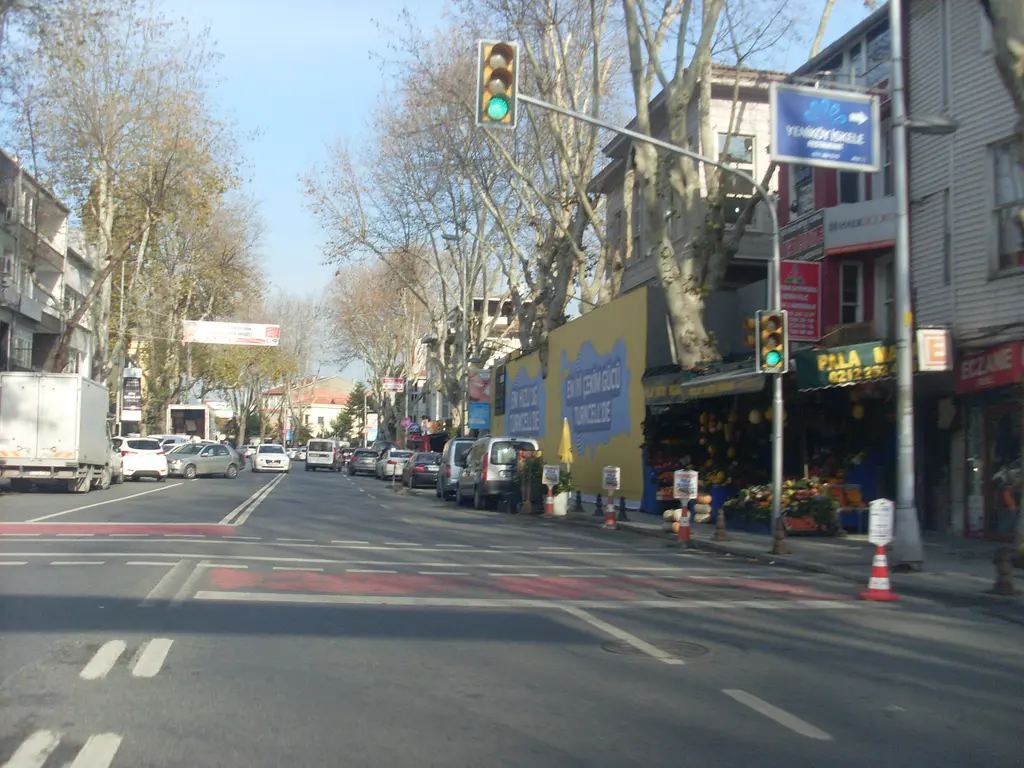 Yeniköy çarşısı; trafik ışıkları, manav ve dükkânlar, cadde boyunca çınarlar ve park etmiş arabalar.