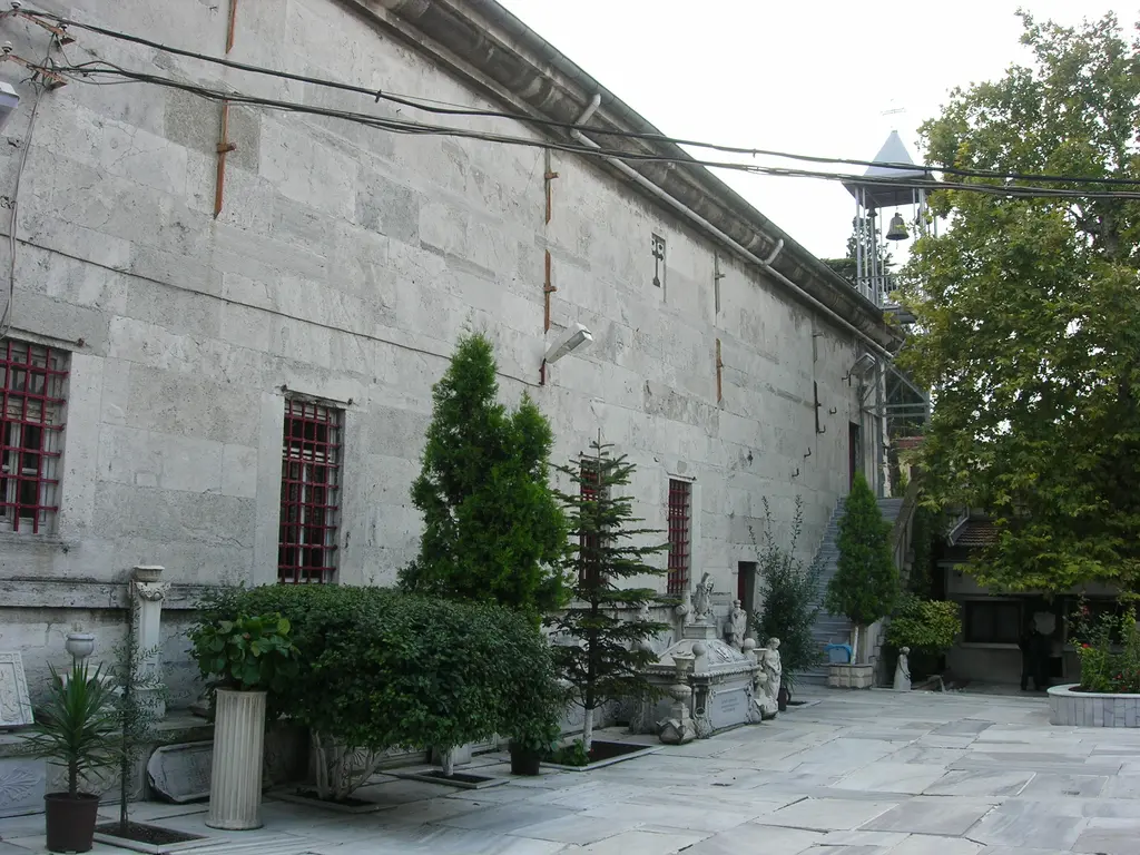 Manastırın avlusundan kilisenin yan cephesi; kesme taş duvar, kırmızı çerçeveli pencereler, sağda metal çan kulesi ve avluda servi ile çalılar.
