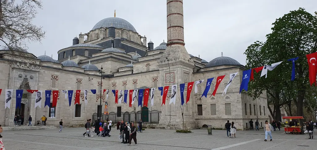 Beyazıt Meydanı'ndan cami; sıra sıra asılmış bayrakların altında kubbeler ve iki minare, önde ağaçlar ve meydanda dolaşan insanlar.