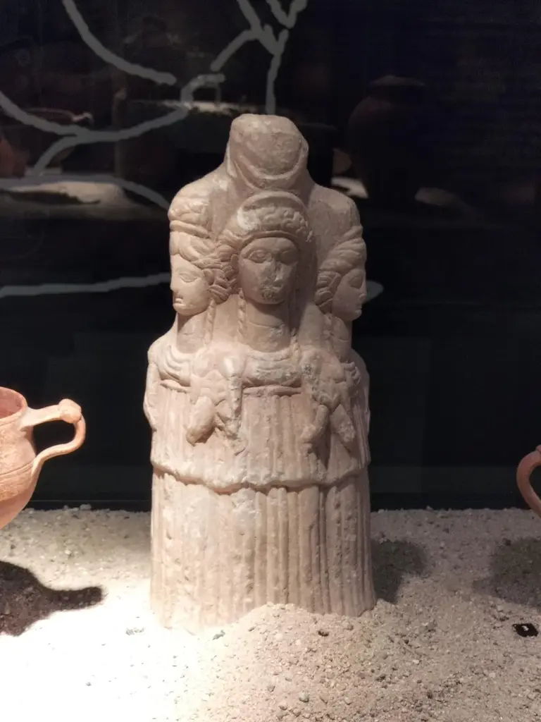 Üçlü Hekate heykelciği; kum zemin üstünde sırt sırta duran üç kadın figürlü taş heykelcik ve iki yanında pişmiş toprak kaplar.