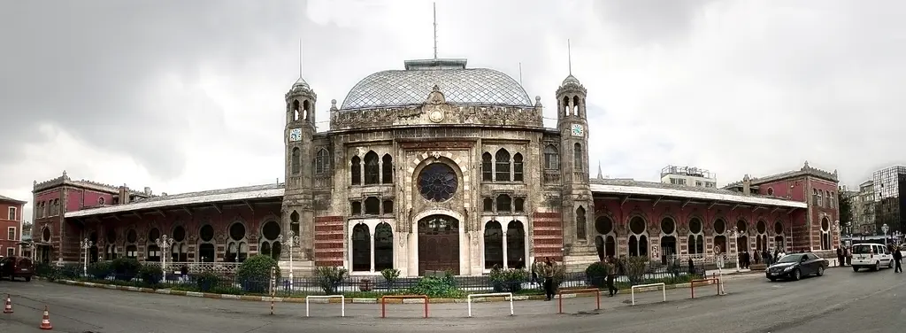 Sirkeci Garı'nın bütün cephesi tek karede; ortada kubbeli giriş bloğu ile iki kule, iki yana uzanan kemerli pencereli kanatlar, önde alçak çit ve kırmızı beyaz korkuluk.