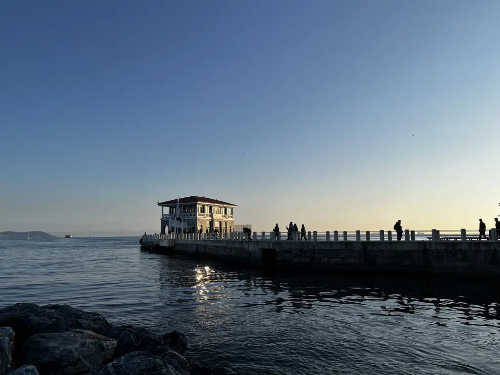 Moda İskelesi; alacakaranlıkta siluet hâlinde bina, iskele yolunda insanlar ve önde kayalar.