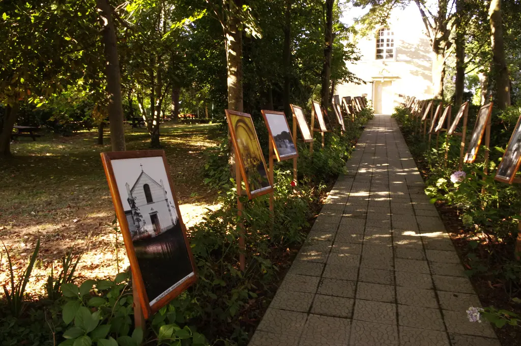 Polonezköy'de ağaçlı bahçenin içinden geçen taş döşeli yürüyüş yolu; iki yanda çerçeveli eski fotoğraf panoları ve ilerde beyaz bir yapı.