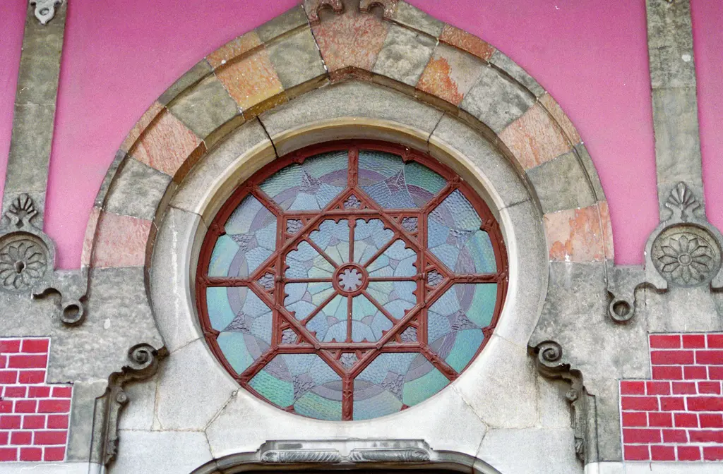 Sirkeci Garı'nda bir gül pencere yakın planda; pembe duvarda sekiz kollu yıldız biçiminde kırmızı demir kafes, turkuaz mavi ve kehribar camlar, çevresinde renkli mermer kemer.