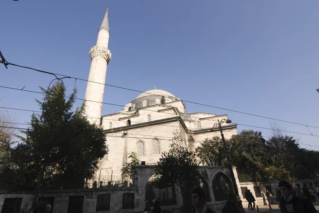Yeniçeriler Caddesi'nden Gazi Atik Ali Paşa Camii; tramvay telleri, avlu duvarı ve ağaçlar ardında kubbe ile ince minare.