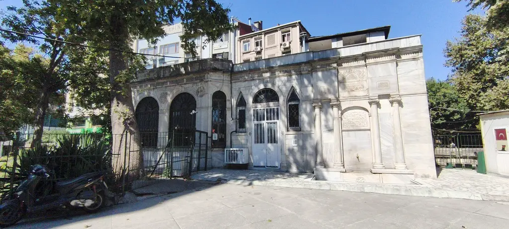 Murat Paşa Camii avlusunda Oğlanlar Tekkesi türbesi; mermer cephe, kemerli pencereler, yivli kuşak ve çevresinde ağaçlar.