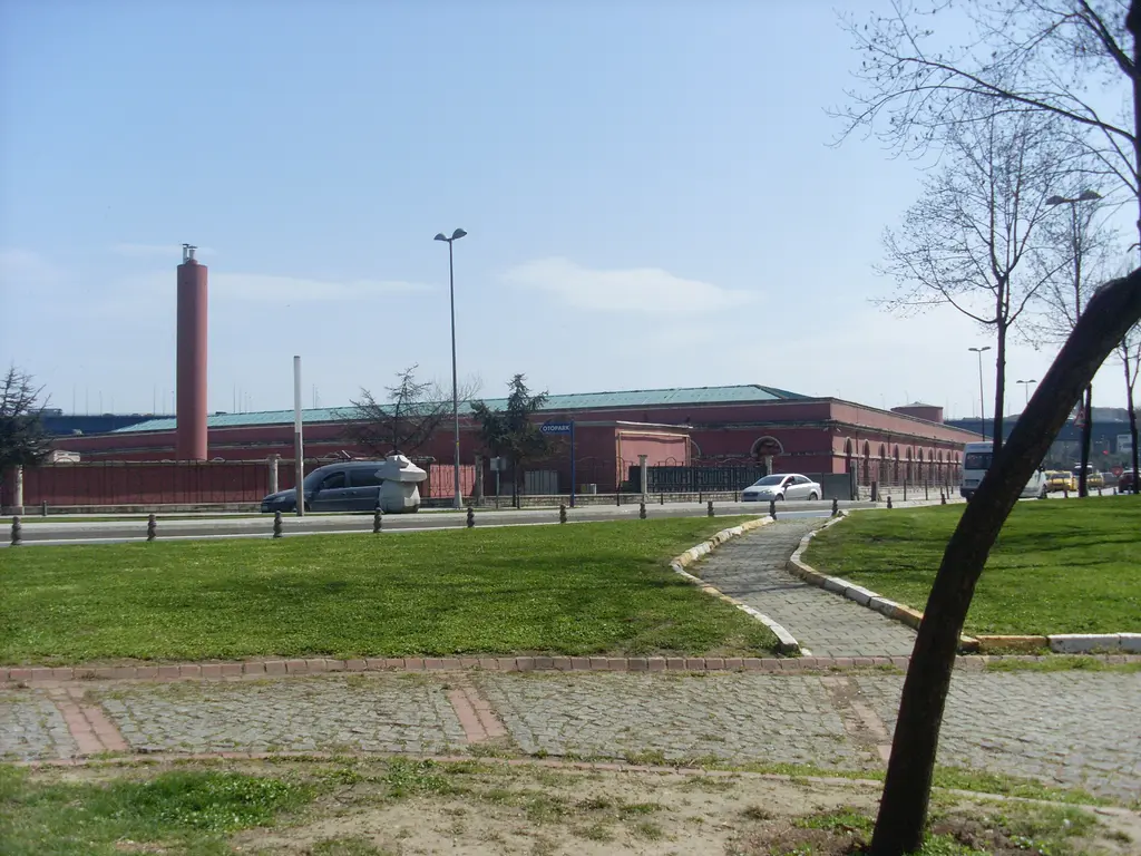 Feshane'nin uzaktan görünümü; kıyıdaki parkın çimenleri ve yolları, arkada uzun tuğla bina ve yüksek baca.