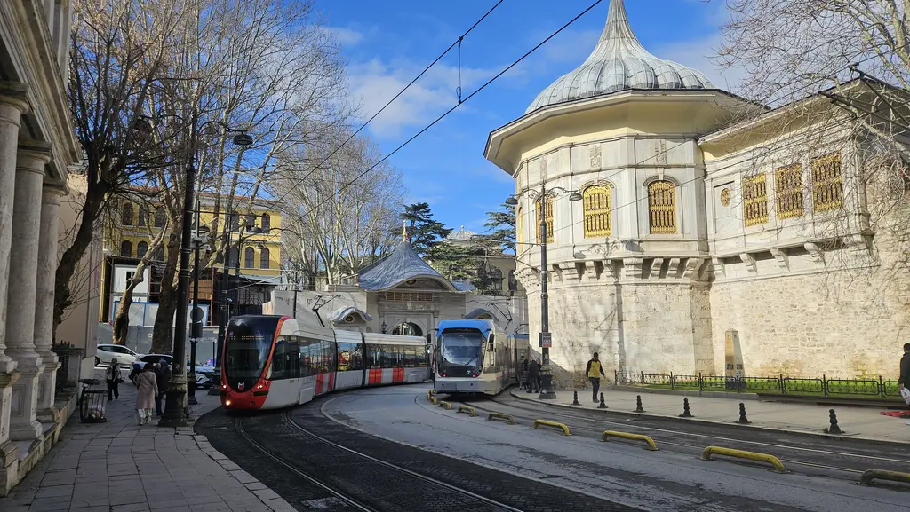 Alay Köşkü önünden geçen iki tramvay; burç üstünde beyaz köşk, soğan kubbe, sur boyunca kafesli pencereler ve yapraksız ağaçlar.