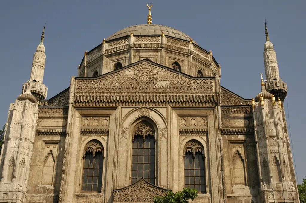 Pertevniyal Valide Sultan Camii'nin güneydoğu cephesi; altın rengi ışık alan taş yüzey, gotik pencereler, üçgen alınlık ve minareler.