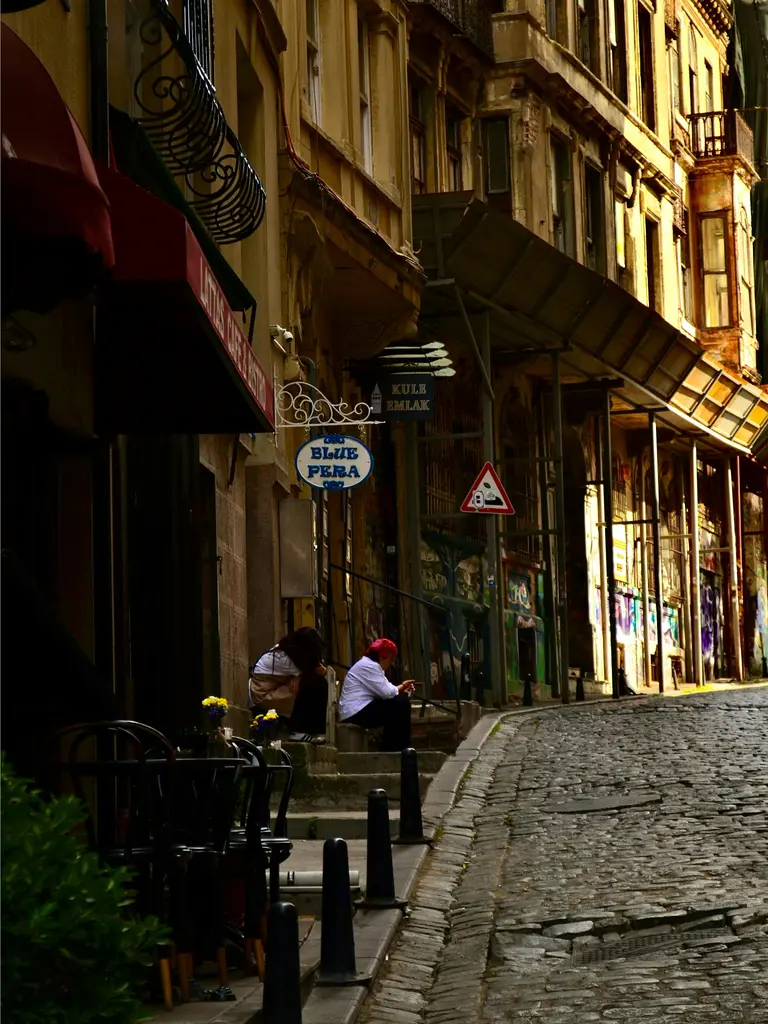 Beyoğlu'nda arnavut kaldırımlı bir yokuş; tentelerin altında oturan insanlar, eski apartman cepheleri ve kıvrılan taş yol.
