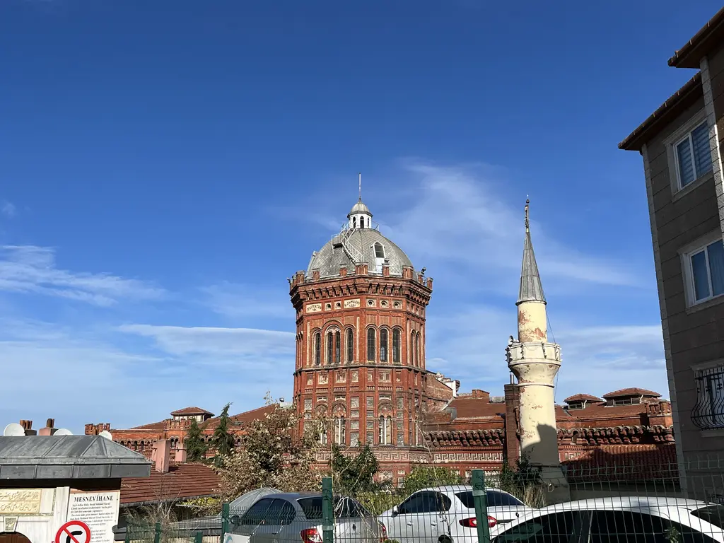 Mahalle sokağından okul; önde apartmanlar ve dükkânlar, arkada ağaçların üstünde okulun kulesi.