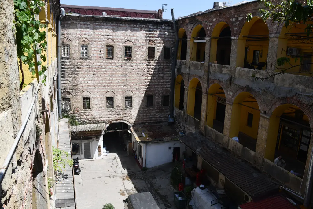 Büyük Yeni Han'ın avlu dibindeki pencereli taş kütle; yanında sarı revaklı kanat, altta beyaz badanalı dükkân sırası.