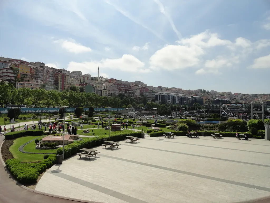 Miniatürk'te taş döşeli meydan; önde ahşap piknik masaları, solda çimenler ve küçük maketler, arkada mavi çevre duvarı ve tepedeki evler.