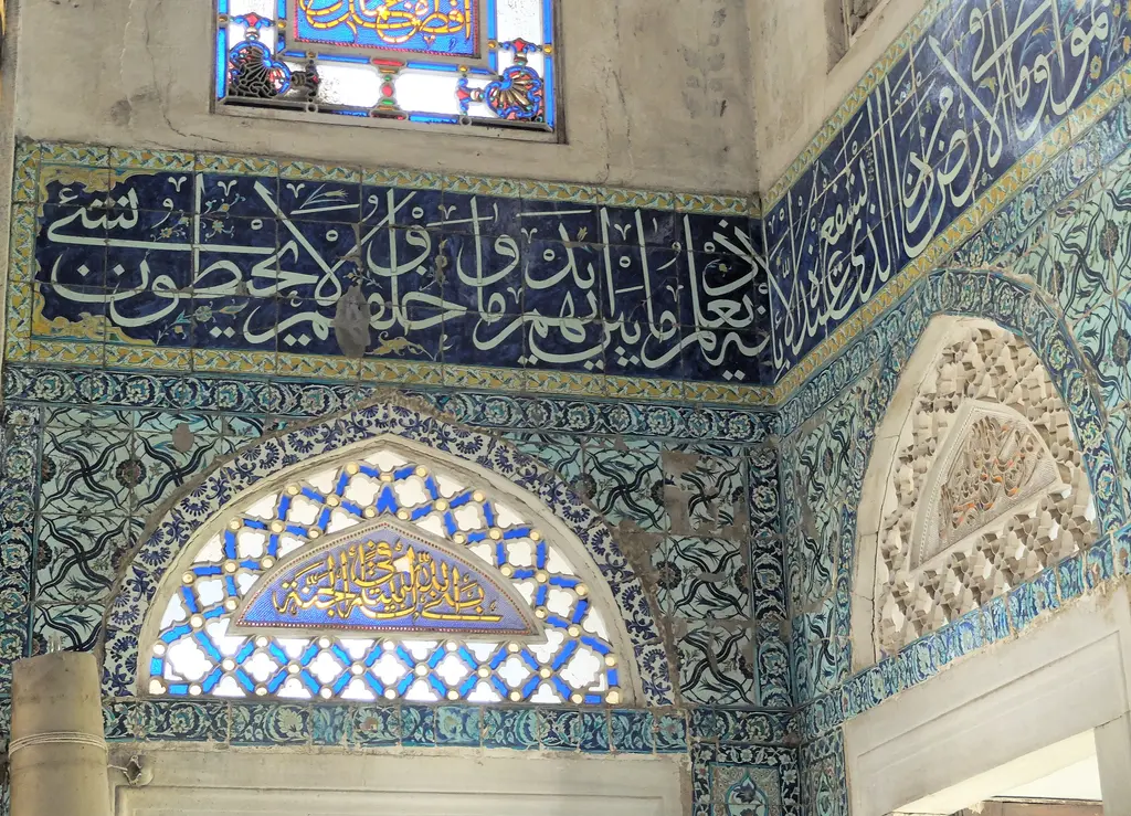 Duvarda çini kaplama; üstte lacivert zemin üzerine beyaz celi sülüs yazı kuşağı, altında firuze bitkisel bordür ve kemerli çini alınlık.