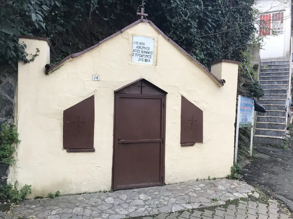 Aya İoannis Ayazması; krem rengi küçük yapı, kahverengi kapı ve panjurlar, çatıda haç ve yanında merdiven.