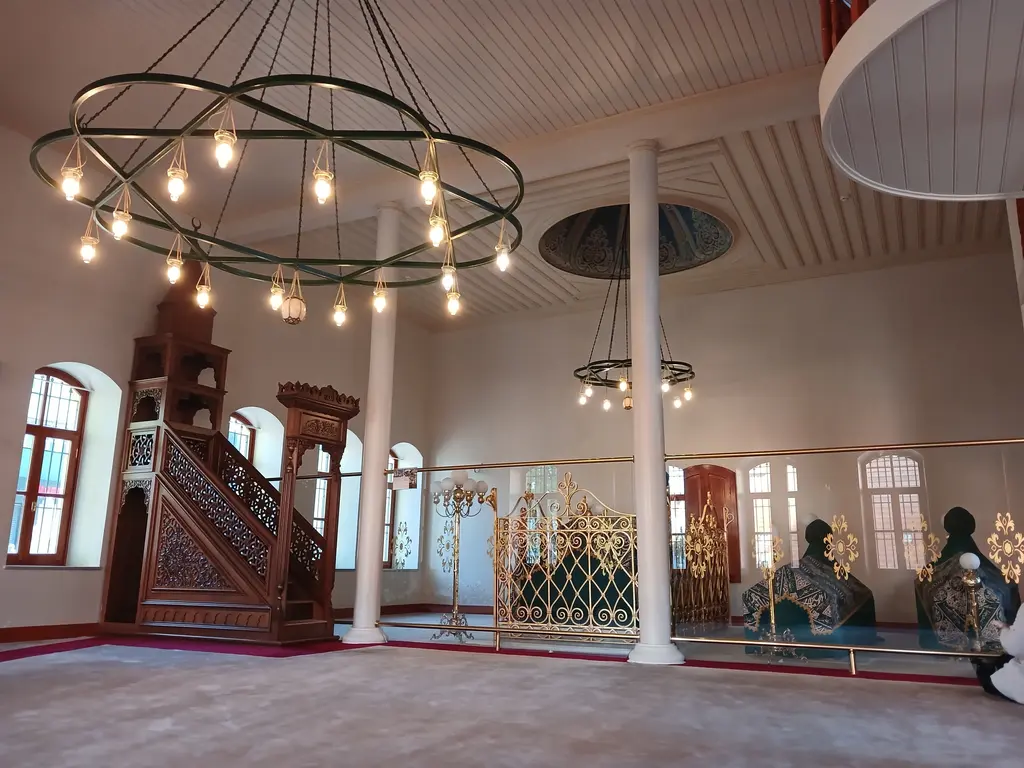Seyyid Nizam Camii'nin içi; beyaz sütunlar, ahşap minber, halka biçimli avize ve sağda parmaklıkla ayrılmış sandukalar.