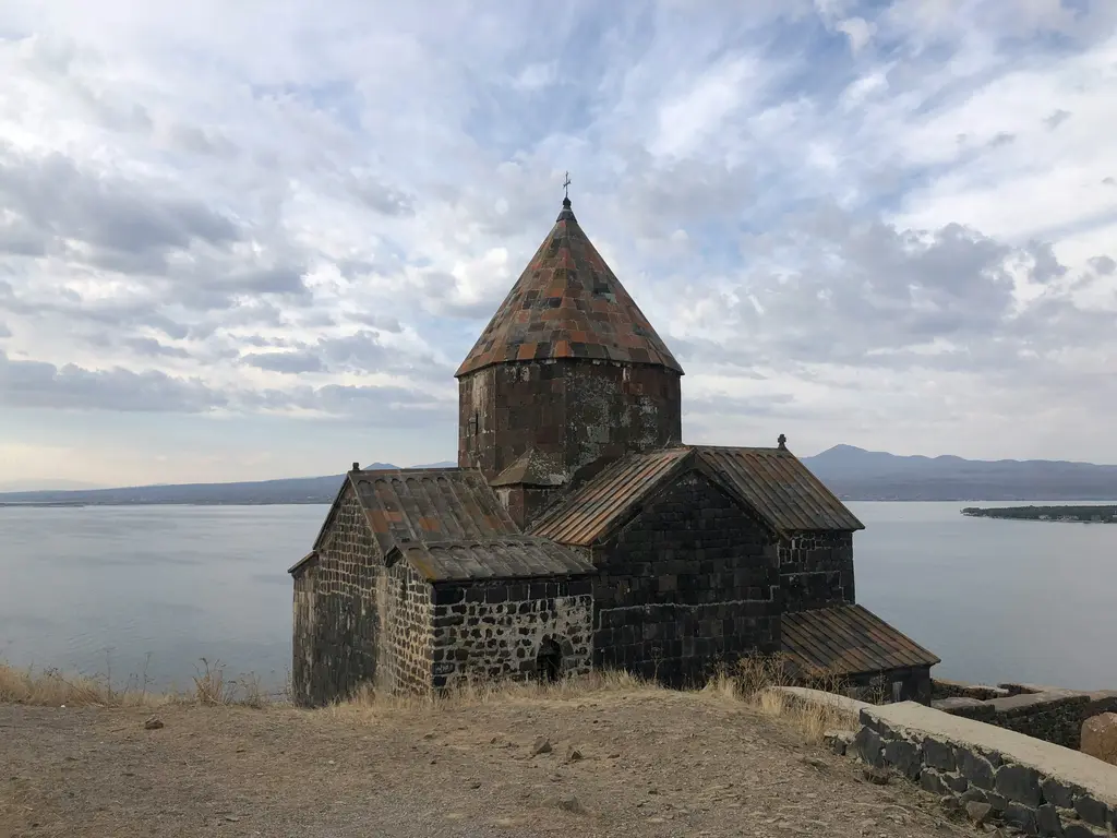 Sevan Gölü'ne bakan tepede duran koyu bazalt taşından kilise; renkli taşlarla örülmüş konik külah, üçgen alınlıklı yan hacimler, gerisinde gölün açık suyu ve karşı kıyıdaki dağlar.