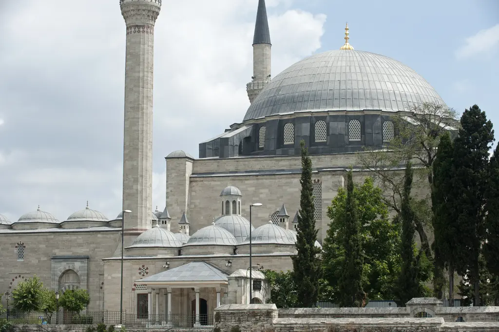 Yavuz Sultan Selim Camii'nin kubbesi yakından; pencereli kasnak, kurşun kaplama, solda kırmızı bantlı minare gövdesi, önde tabhane kubbeleri ve serviler.