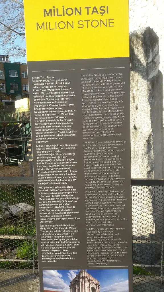 Milyon Taşı'nın yanında sarı başlıklı bilgi panosu; Türkçe ve İngilizce metin, altında bir görsel, arkada tel çit ve mermer ayak.