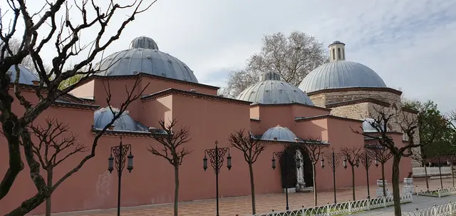 Sultanahmet'te Haseki Hürrem Sultan Hamamı; somon rengi sıvalı uzun duvar, arkasında sıralanan kurşun kaplı kubbeler ve önünde çıplak ağaçlarla demir korkuluk.