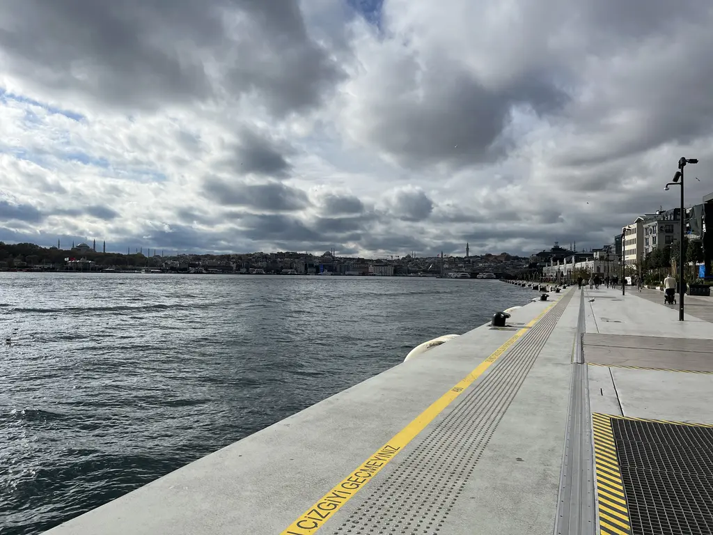 Galataport'ta rıhtım; sağda binalar ve ağaçlar, ortada geniş beton kıyı ve sarı çizgiler, solda Boğaz'ın dalgalı suyu.