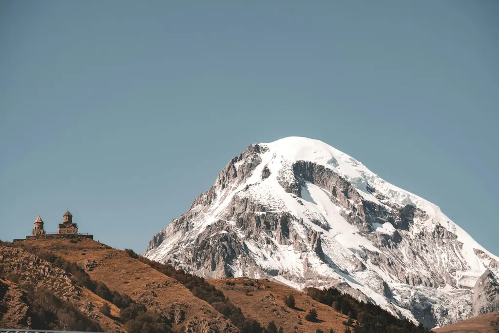 Kazbegi'de tek başına bir tepede duran Gergeti Üçlü Kilisesi; arkasında karlı Kazbek Dağı ve bulutlar.