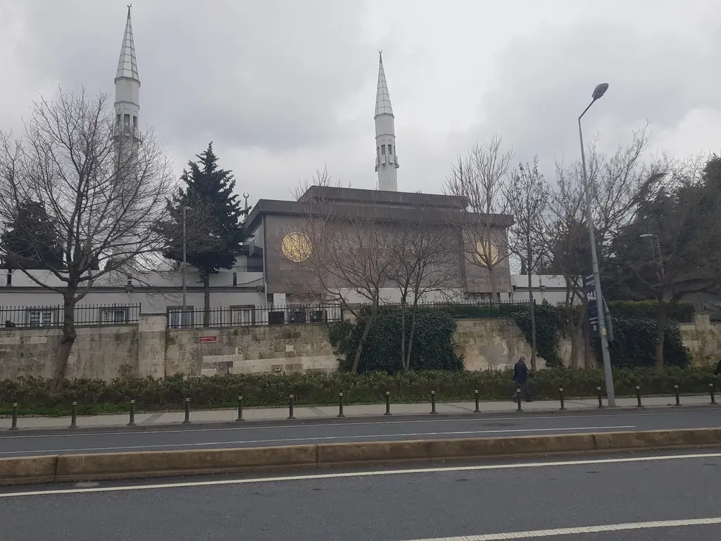 Şakirin Camii'nin caddeden görünümü; taş istinat duvarı, ağaçlar, alçak cami kütlesi ve gri gökyüzüne yükselen iki minare.