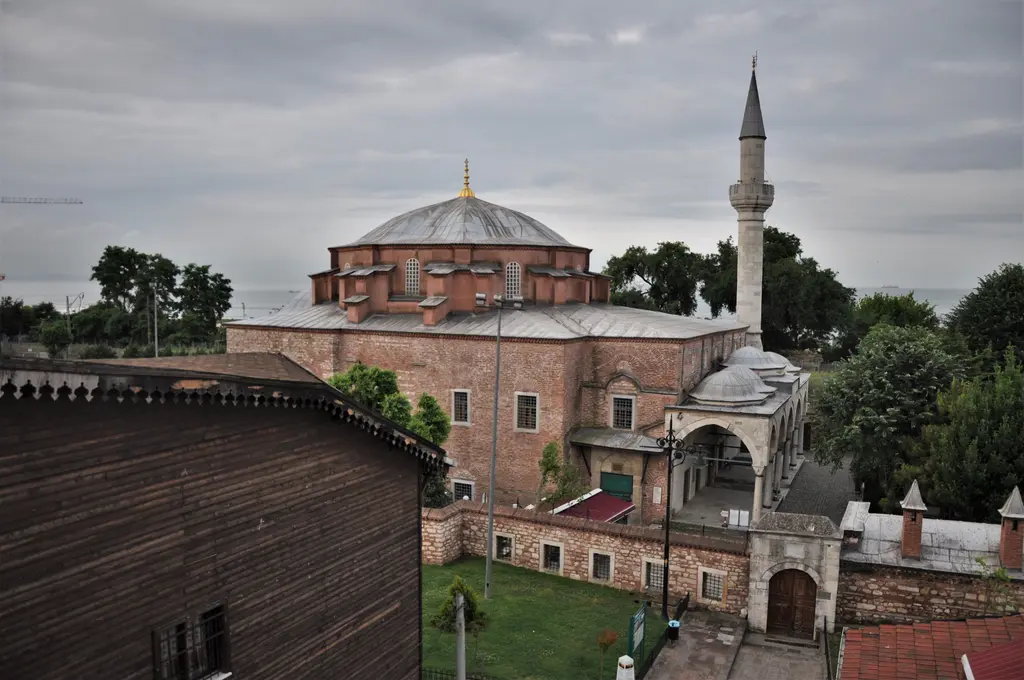 Küçük Ayasofya Camii çatılardan; tuğla taş gövde, kurşun kubbe, sağda tam boy minare, konik külahlı avlu duvarı ve ufukta solda Marmara Denizi'nin şeridi.